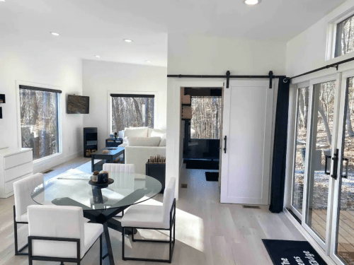 Bright, modern living space with a dining area, large windows, and a view of trees outside. Minimalist decor and furniture.