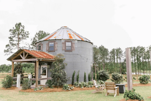 A rustic, round metal building with a rusty roof, surrounded by greenery and a small porch with seating.