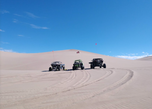 Three off-road vehicles parked on sandy dunes under a clear blue sky, with tire tracks leading into the distance.