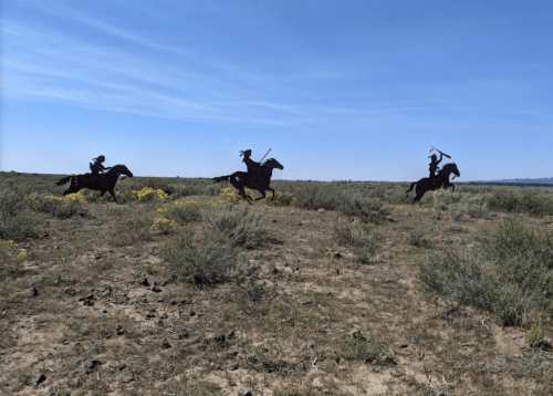 Silhouettes of three horsemen riding through a sparse, grassy landscape under a clear blue sky.