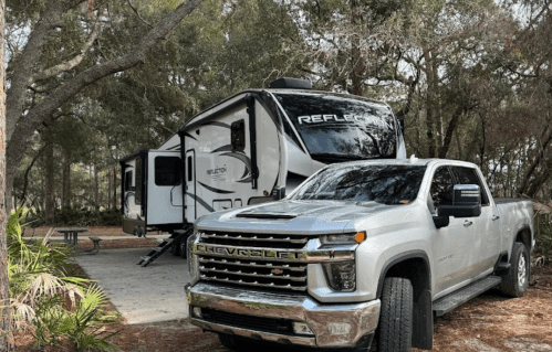 A silver pickup truck parked in front of a large RV at a campsite surrounded by trees.