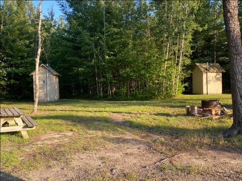 Two small cabins in a wooded area, with a picnic table and fire pit in the foreground on a grassy path.