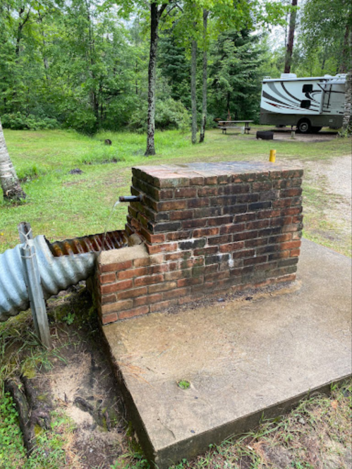 A brick well structure with a metal pipe, surrounded by greenery and a camping area in the background.