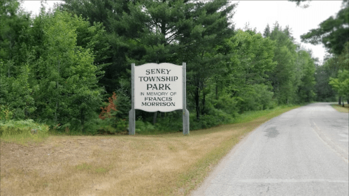 Sign for Seney Township Park in memory of Francis Morrison, surrounded by trees along a gravel road.