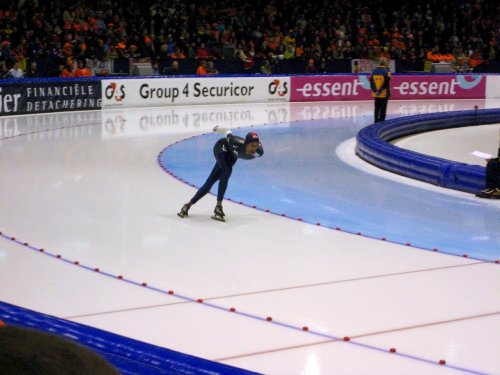 A speed skater leans forward on the ice track, with a cheering crowd in the background.