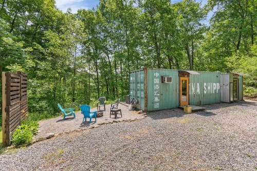 A green shipping container converted into a cozy outdoor space, surrounded by trees and gravel, with chairs arranged outside.