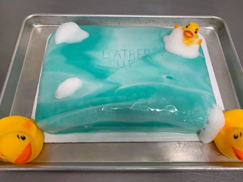 A rectangular cake with blue icing, featuring rubber duck decorations and foam, labeled "LATHER UP."