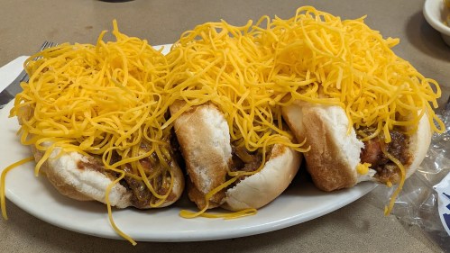 Three hot dogs topped with a generous amount of shredded cheddar cheese on a white plate.