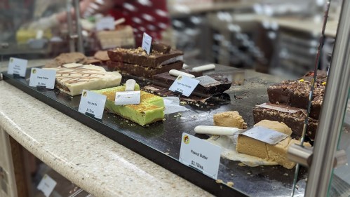 A display of various fudge flavors with labels, including chocolate, peanut butter, and mint, in a glass case.