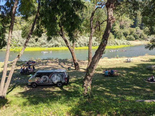 A scenic riverside park with people picnicking, a colorful food truck, and kayakers on the water.