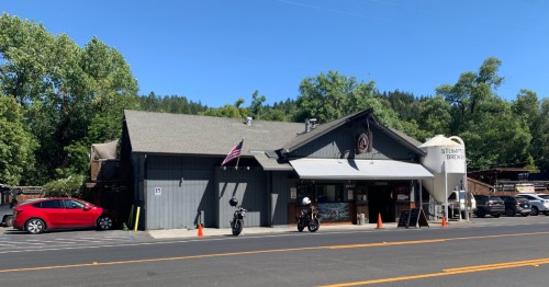 A gray building with an American flag, surrounded by trees, featuring a brewery sign and parked motorcycles.