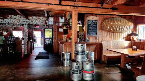 Interior of a brewery featuring wooden decor, beer taps, and stacked kegs, with patrons in the background.