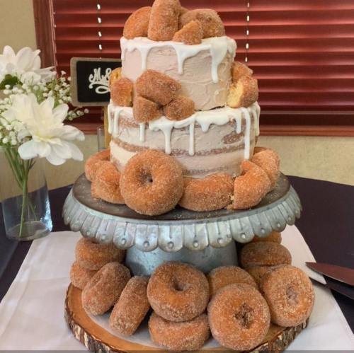 A tiered cake topped with donuts, surrounded by more donuts on a rustic stand, with flowers in the background.