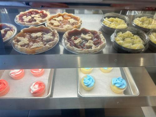 A display case with fruit pies, plastic containers of yellow dessert, and frosted cupcakes in pink and blue.
