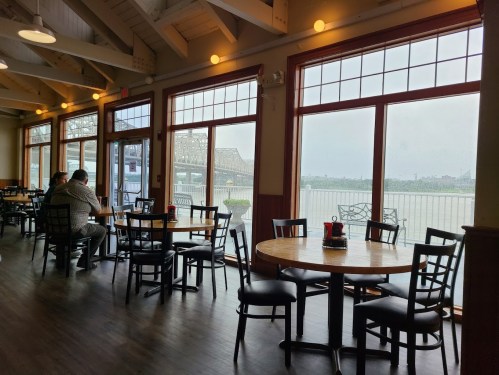 Interior of a restaurant with wooden tables and chairs, large windows overlooking a river and bridge. Two patrons seated.