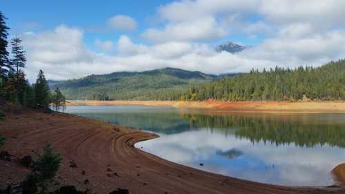 A serene lake surrounded by trees and mountains, reflecting clouds and blue sky on calm water.