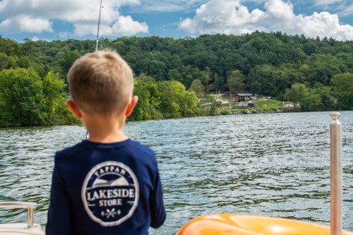 A child in a blue shirt gazes at a serene lake surrounded by green trees and a distant lakeside store.