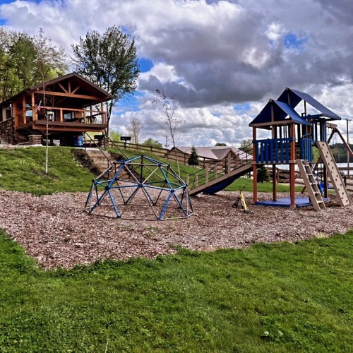 A playground with a climbing structure and slide, surrounded by grassy areas and trees under a cloudy sky.