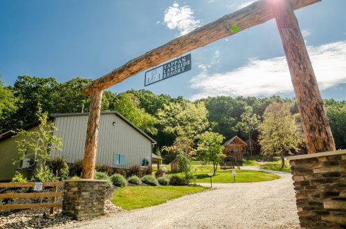 A rustic entrance sign welcomes visitors to a lodge resort, surrounded by greenery and blue skies.