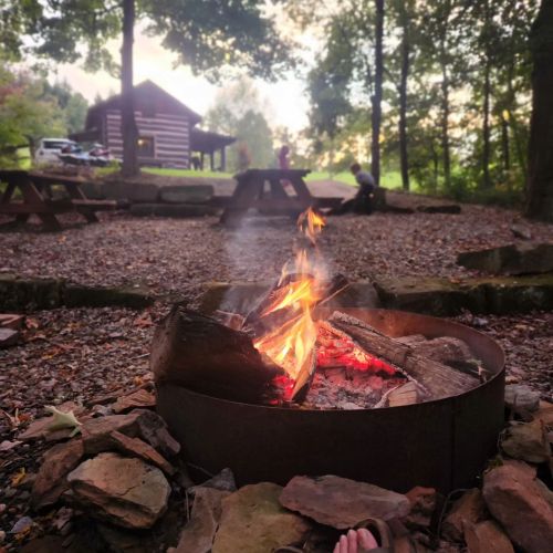 A campfire burns in a stone ring, with a log cabin and picnic tables in the background, surrounded by trees.