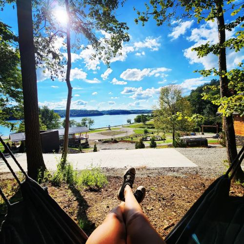 A person relaxes in a hammock with a scenic view of a lake and trees under a bright blue sky.