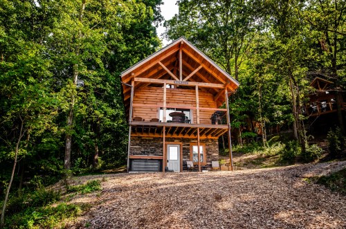 A wooden cabin with a sloped roof, surrounded by trees, set on a gravel path in a forested area.