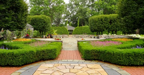 A manicured botanical garden featuring circular stone paths, trimmed hedges, and a white gazebo.