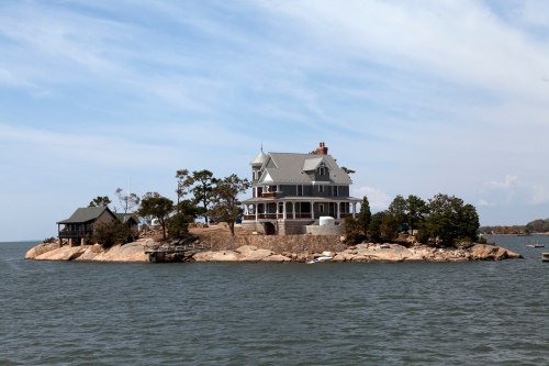 A large, elegant house on a small island surrounded by water, with trees and rocky shoreline in the background.