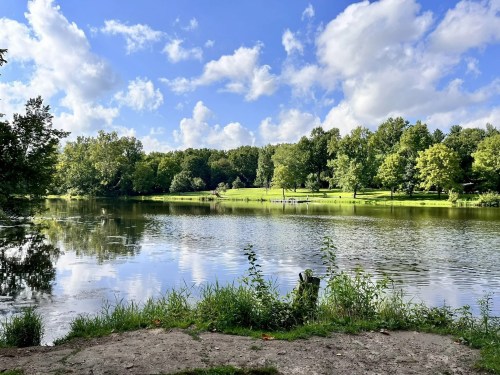 A serene lake surrounded by lush green trees and a blue sky with fluffy clouds reflecting on the water's surface.