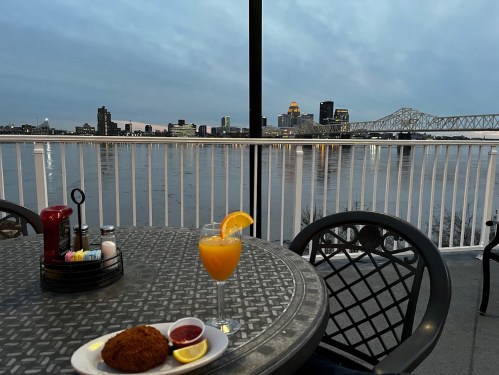 A table with a drink and appetizer overlooks a river and city skyline at dusk.