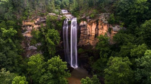 A stunning waterfall cascades down rocky cliffs, surrounded by lush green trees in a serene natural setting.