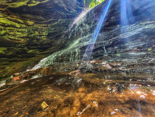 A serene waterfall cascades over rocky terrain, surrounded by lush greenery and illuminated by sunlight beams.