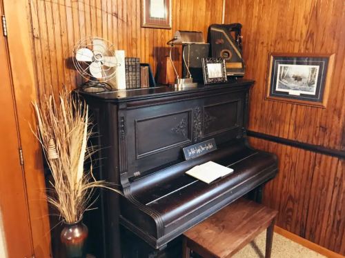 A vintage piano in a cozy room, surrounded by decor like books, a fan, and framed photos against wooden walls.