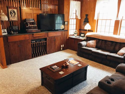 Cozy living room with wooden paneling, a TV, a coffee table, and comfortable seating. Natural light from windows.