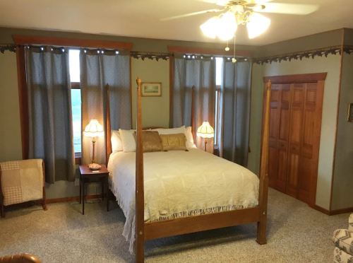 Cozy bedroom featuring a four-poster bed, two lamps, and large windows with curtains. Warm, inviting decor.