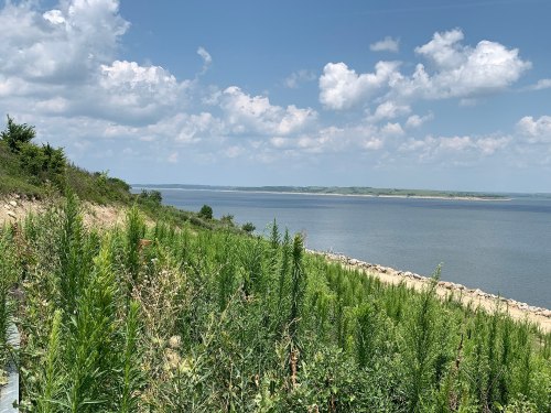 A scenic view of a lake surrounded by lush greenery and blue skies with scattered clouds.