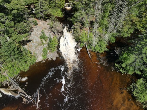 Aerial view of a waterfall cascading into a dark river, surrounded by lush green trees and rocky terrain.