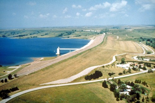 Aerial view of a dam with a reservoir, surrounded by grassy land and a winding road. Clear blue sky above.