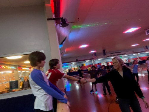 A woman reaches out to two boys at a roller skating rink, with colorful lights and other skaters in the background.