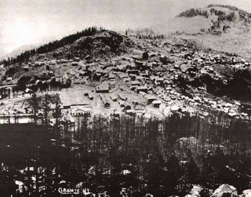 Aerial view of a historic mining town nestled in a mountainous landscape, surrounded by trees and rocky terrain.