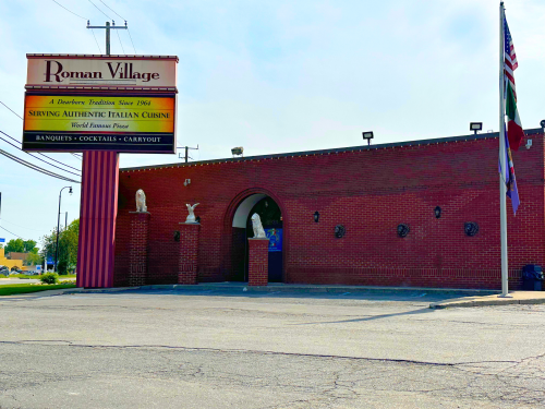 Exterior of Roman Village restaurant, featuring a red brick facade, sign, and flags, with a clear blue sky above.