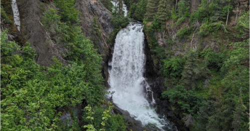 A cascading waterfall surrounded by lush green trees and rocky terrain.
