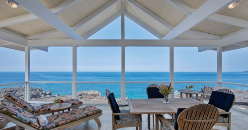 Cozy seaside patio with lounge chairs and a dining table, overlooking a calm blue ocean and rocky shoreline.