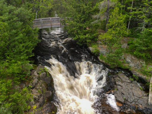 A rushing waterfall flows over rocks beneath a wooden bridge, surrounded by lush green trees.