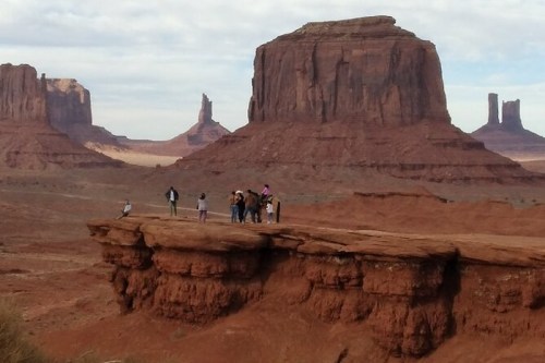 A group of people stands on a rocky ledge in a desert landscape with towering red rock formations in the background.