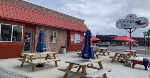 Exterior of Fat Racks BBQ with picnic tables and umbrellas, featuring a red roof and a sign with Texas imagery.