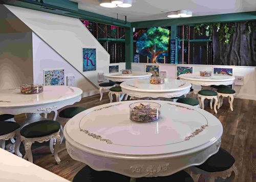 Brightly lit room with elegant round tables, each featuring a centerpiece, surrounded by stylish black stools.