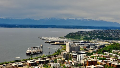 Aerial view of a waterfront city with mountains in the background, featuring a harbor and urban buildings.
