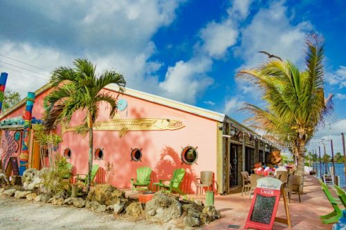 Colorful waterfront building with palm trees, outdoor seating, and a mural of a fish against a blue sky.