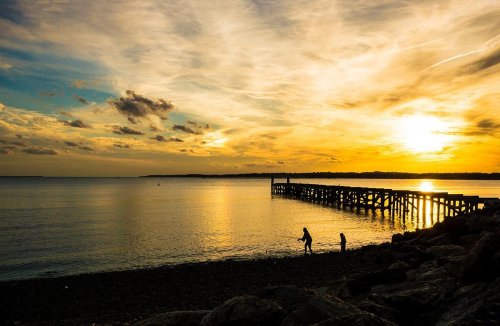Silhouetted figures walk along a rocky shore at sunset, with a pier extending into the calm water.